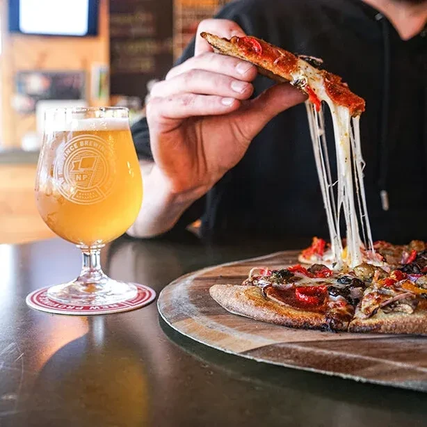 Hand holds pizza slice with stretching cheese next to Prince Brewing beer.
