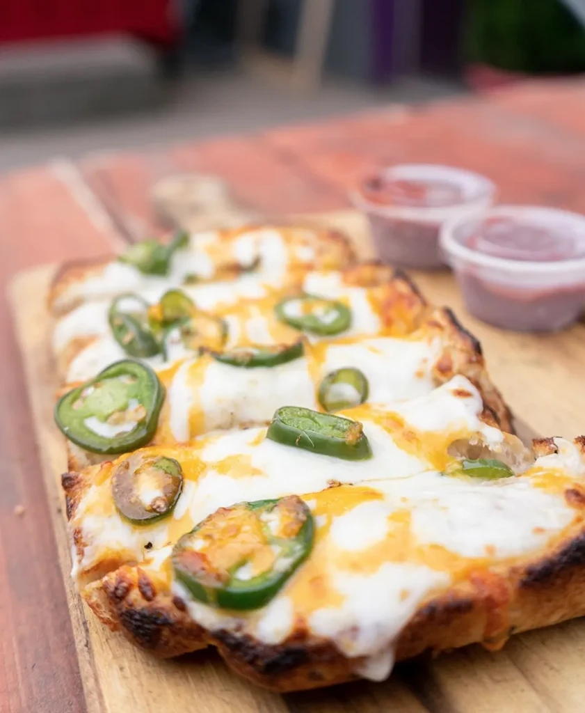 Rectangular cheesy jalapeño flatbread with sauce on a wooden board.