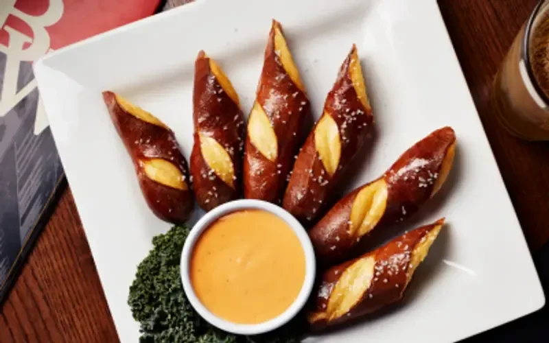 Six golden pretzel sticks with orange dip and greens.