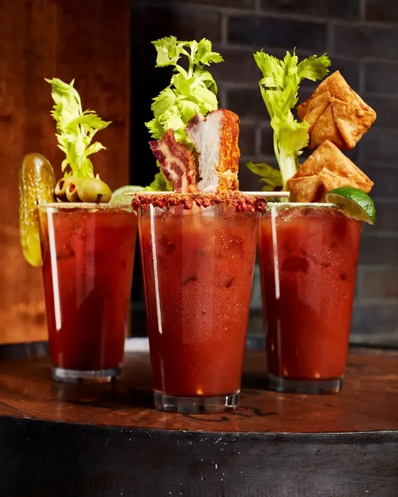 Three elaborately garnished Bloody Mary cocktails on a rustic wooden surface.