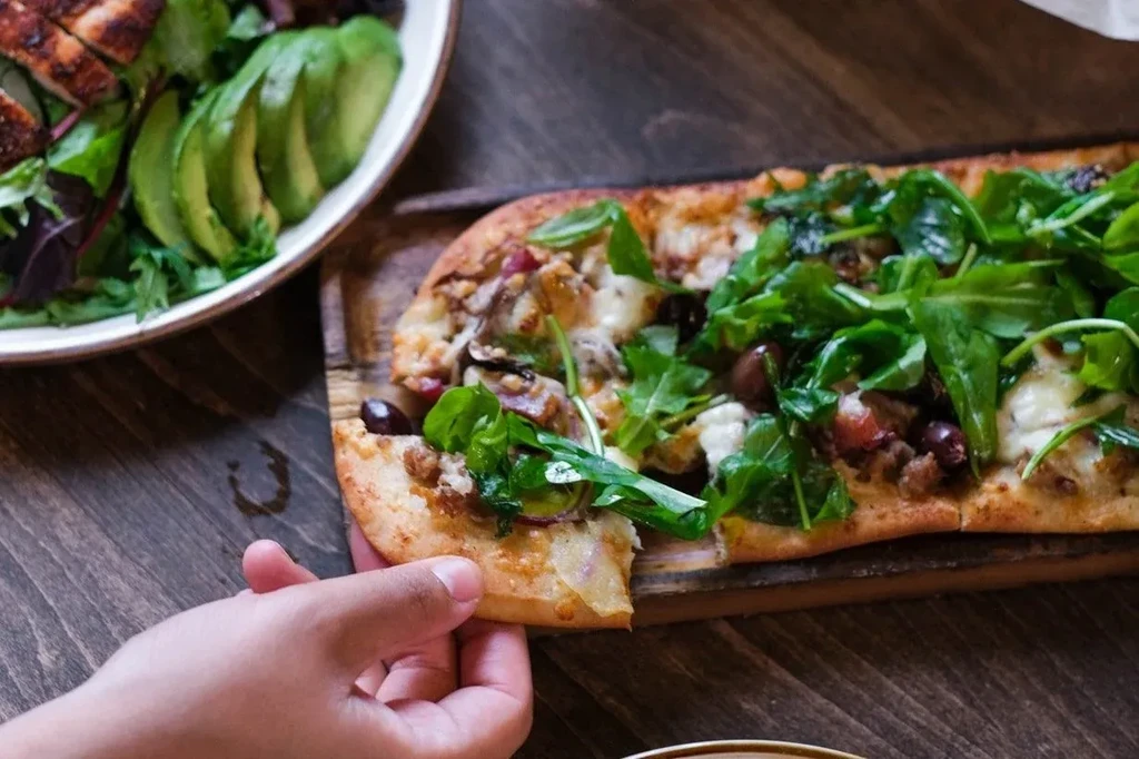 Hand holding flatbread pizza with greens, olives, beside a chicken salad.
