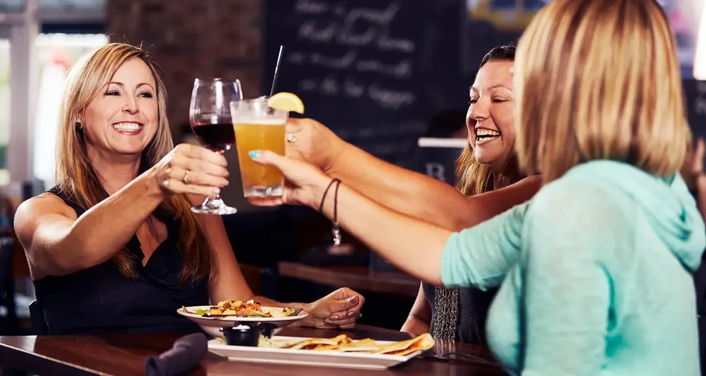 Three smiling women toast with drinks and appetizers in a restaurant.