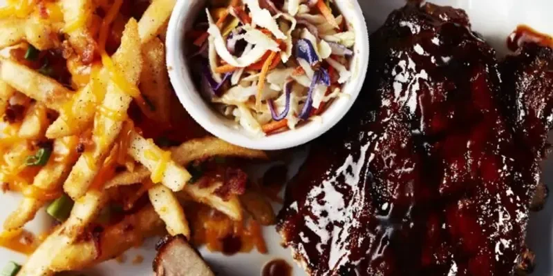 Close-up of rack of BBQ ribs, cheesy fries, coleslaw on white plate.