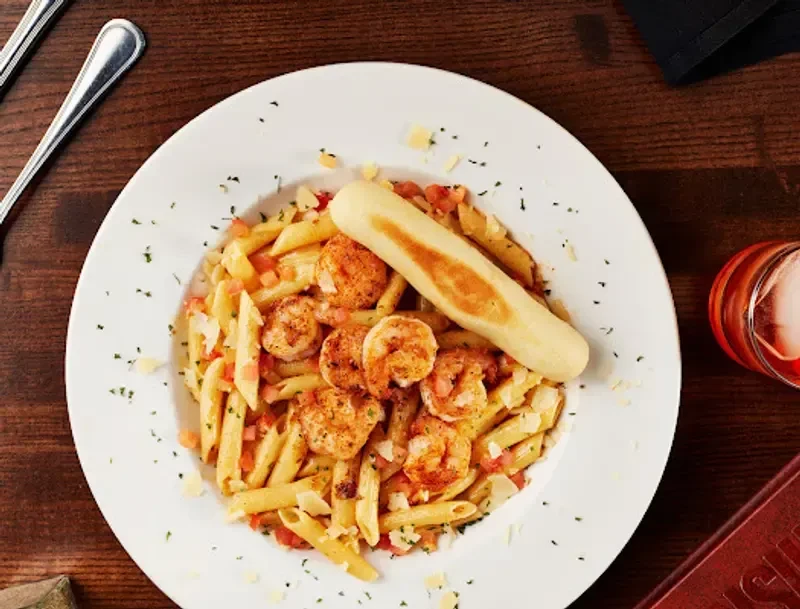 Top-down: Penne pasta with shrimp, breadstick, drink on dark table.