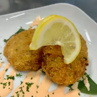 Two golden crab cakes with lemon, sauce, and herbs on a white plate.