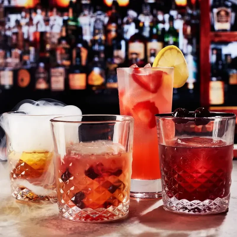 Four colorful, garnished cocktails on a bar surface, one smoking.