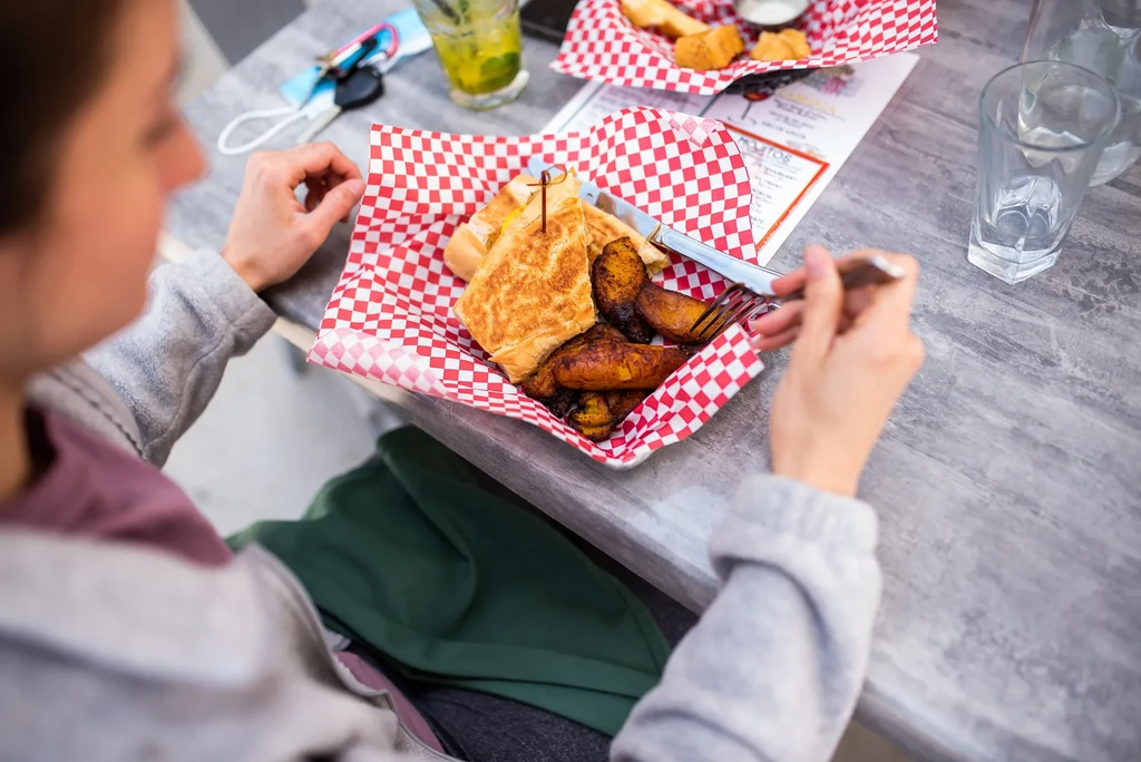 Diner holding cutlery over pressed sandwich, plantains, and a green drink.