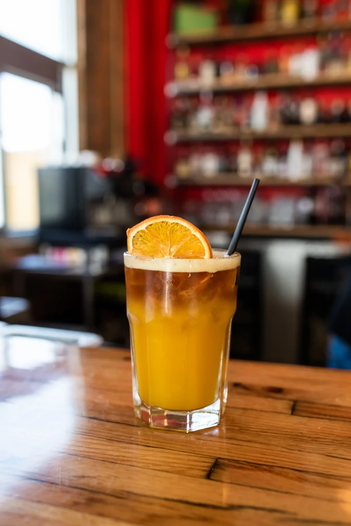 Layered cocktail with orange slice and straw on a wooden bar.