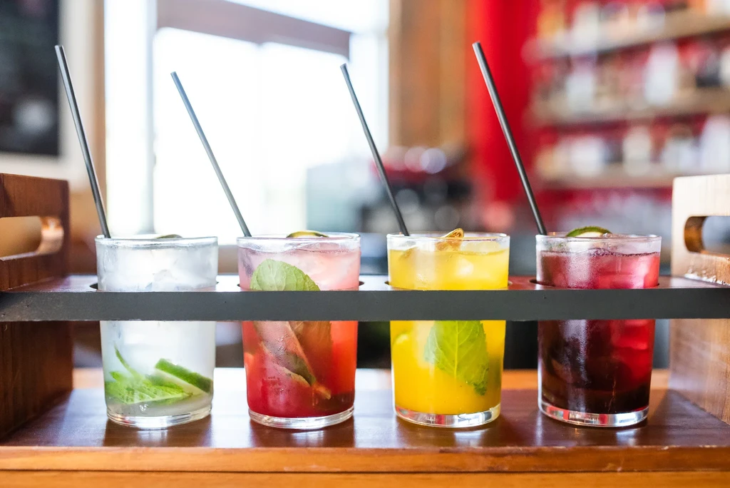 Four colorful garnished drinks in a dark tray on a wooden surface.