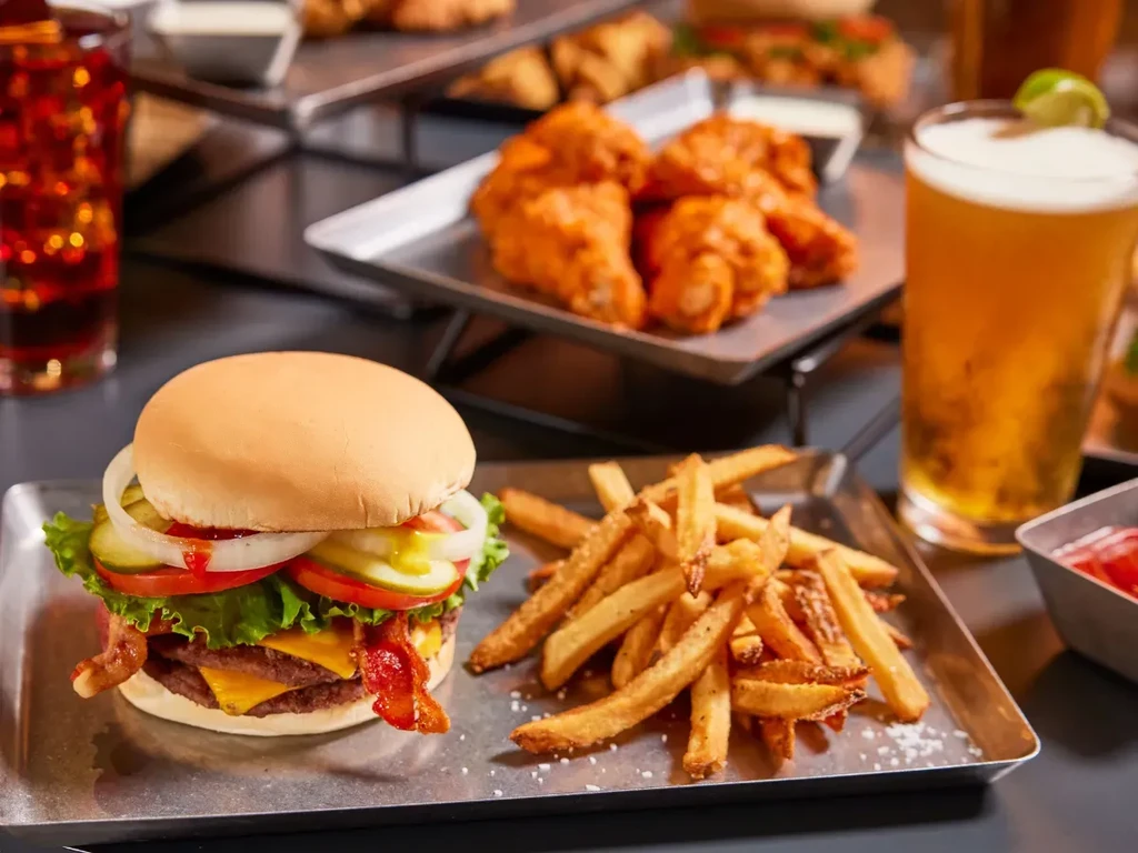 Double cheeseburger, fries, wings, soda, and beer on two metallic trays.