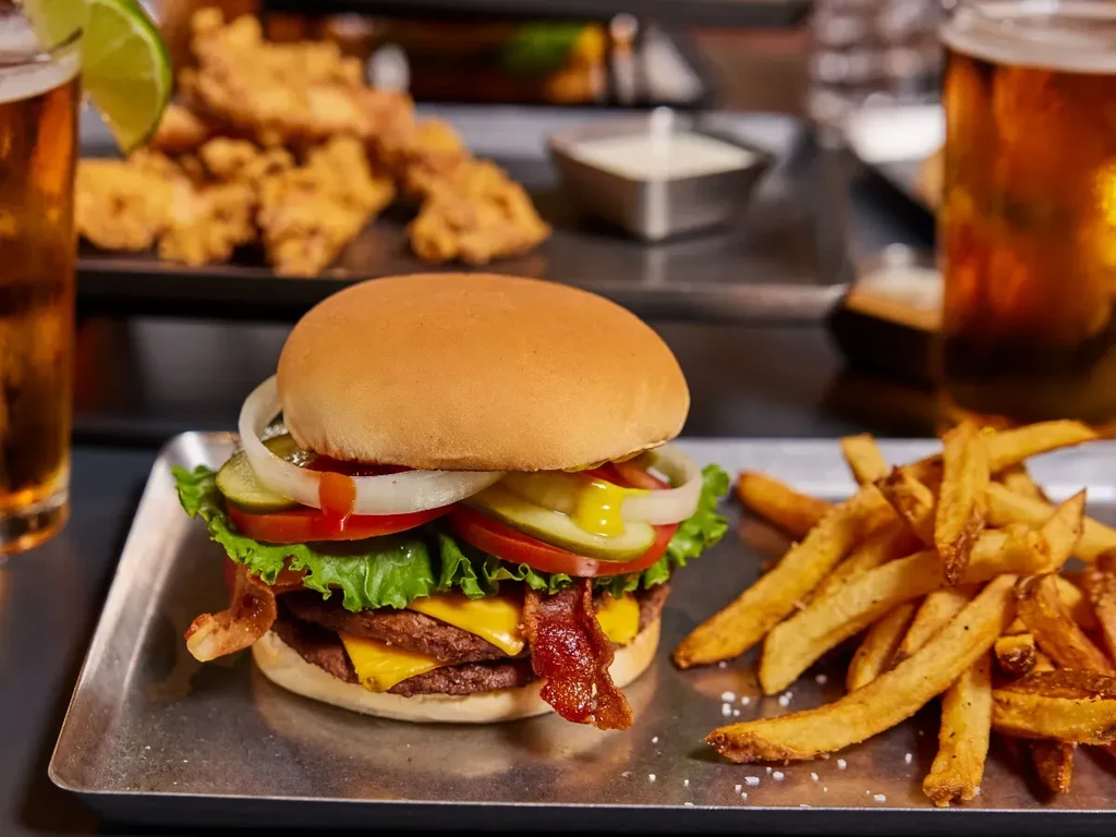 Close-up: Double cheeseburger, fries, beer, fried items on metal trays.