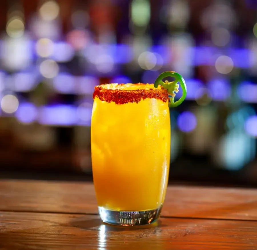 Yellow-orange cocktail, spice-rimmed glass, jalapeño garnish on wood.