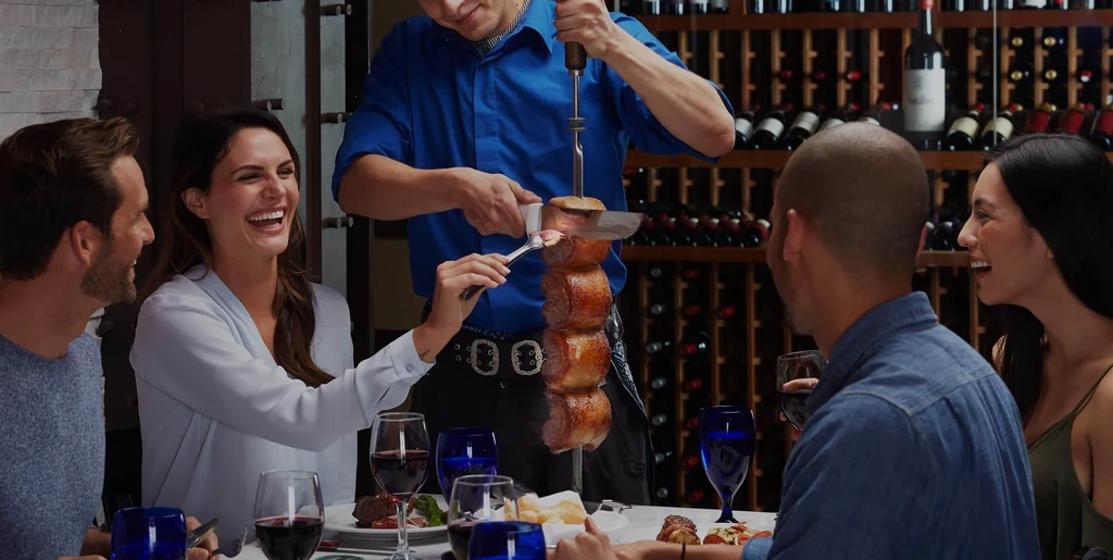 Four smiling diners served skewered meat by a waiter in a restaurant.