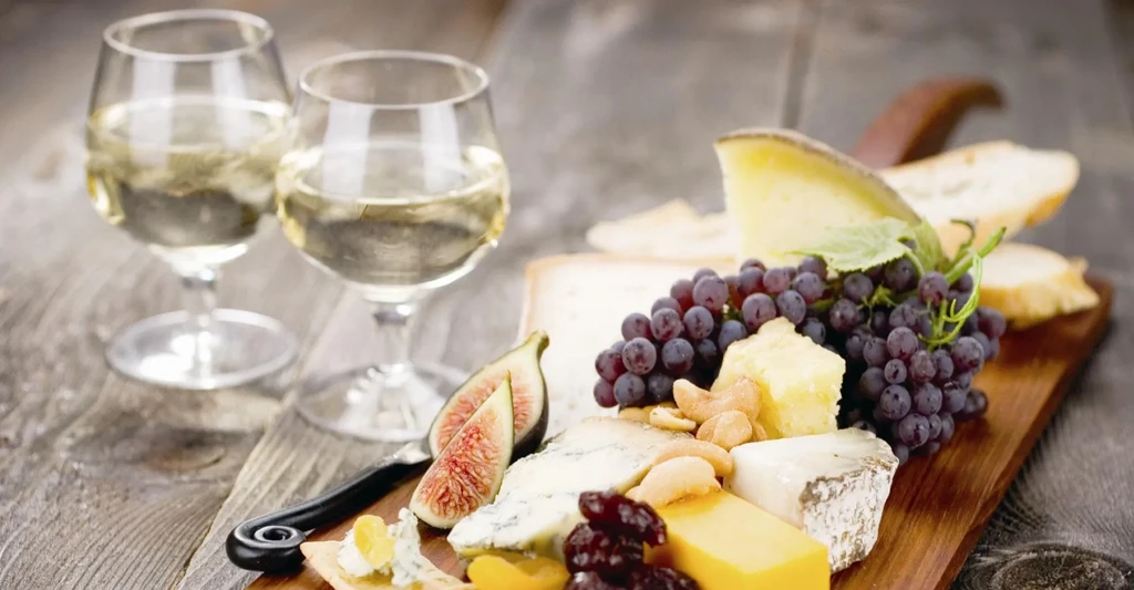 Rustic wooden cutting board with cheeses, fruits, nuts, and wine.