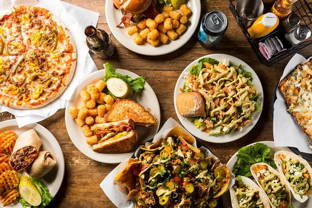 Rustic table with pizza, burgers, nachos, tacos, salad, and drinks.