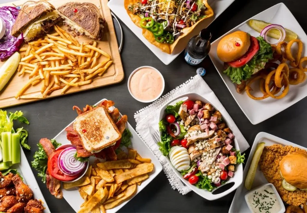 Overhead shot of various comfort food plates, sandwiches, salads on dark table.