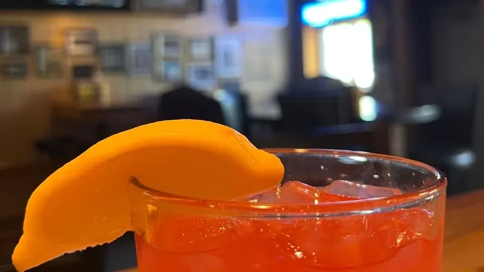 Reddish-orange cocktail with ice and orange slice garnish in a dim bar.