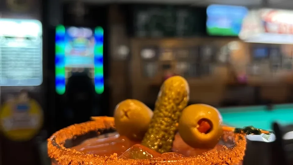 Close-up of a Bloody Mary with pickle, olives, in blurred bar setting.