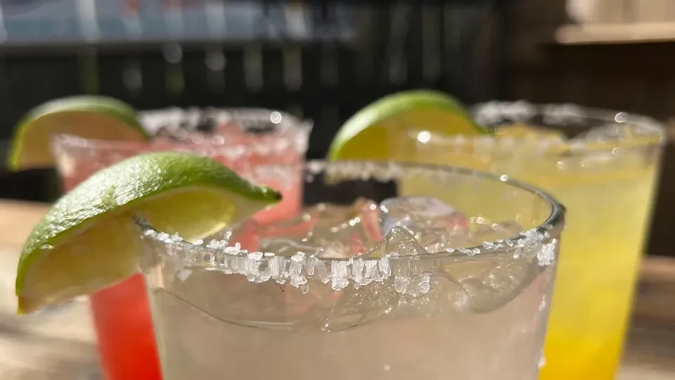 Three colorful cocktails with lime wedges and salted rims on wood.