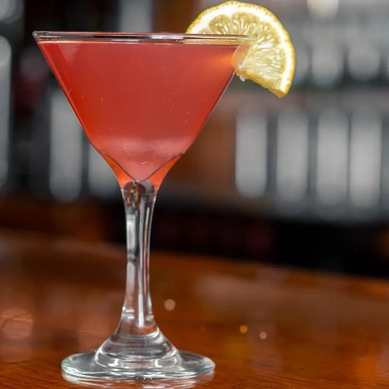 Red-pink cocktail in martini glass with citrus slice on a bar.