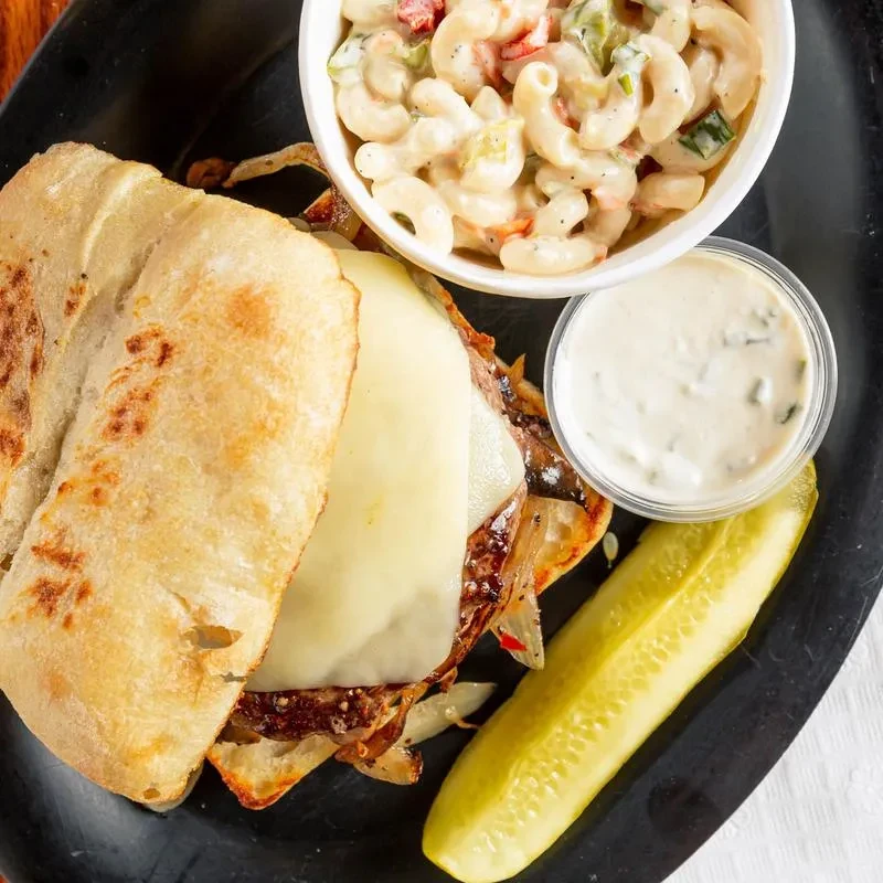 A cheesesteak sandwich, macaroni salad, dipping sauce, and pickle on a dark plate.