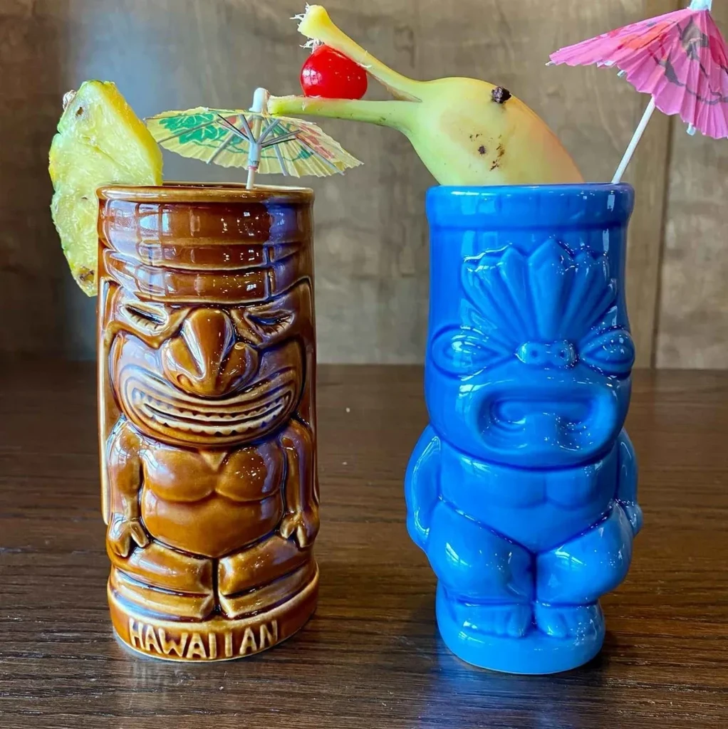 Two tiki mugs, brown and blue, garnished with pineapple, banana, cherry.
