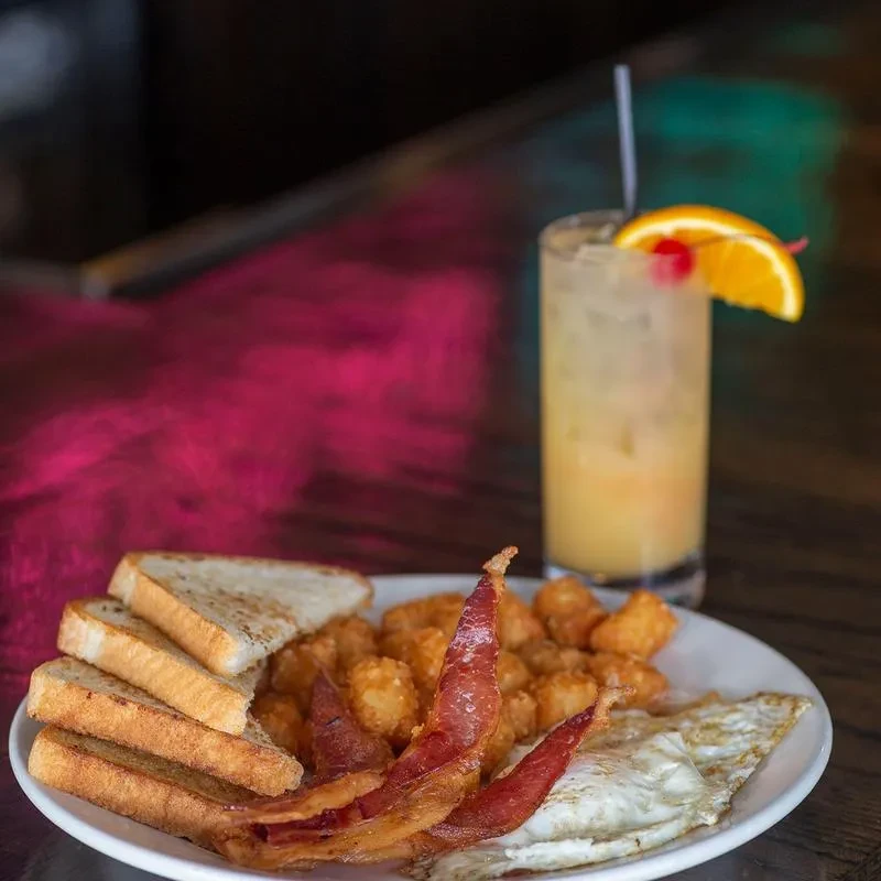Plate of fried eggs, crispy bacon, tater tots, toast, and a cocktail.