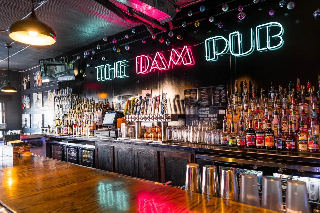 Inside "THE DAM PUB" bar with liquor bottles, beer taps, neon sign.