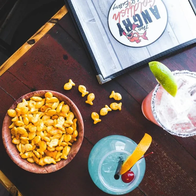 Goldfish crackers, two colorful cocktails, and 'The Angry Goldfish Eatery' logo.