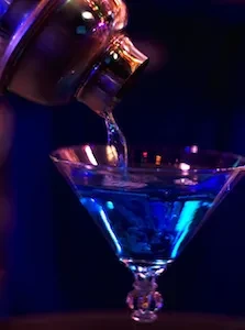 Metallic cocktail shaker pours glowing blue liquid into a martini glass.