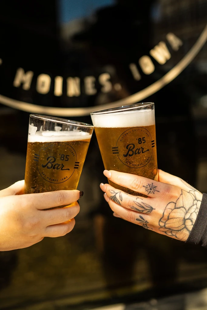Hands clinking "Historic East Village '85 Bar" beer glasses.
