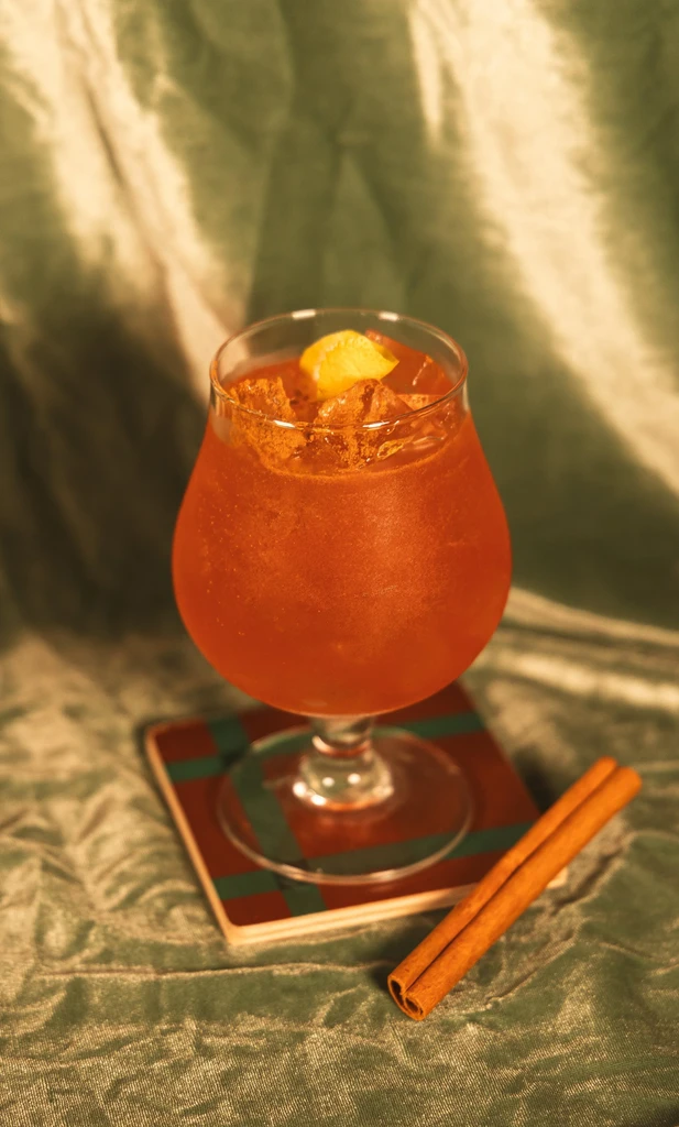 Red-orange cocktail in stemmed glass with ice, orange peel, cinnamon sticks.