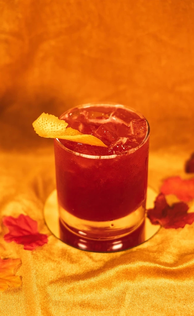 Red cocktail with citrus peel, ice, and autumn leaves on velvet.
