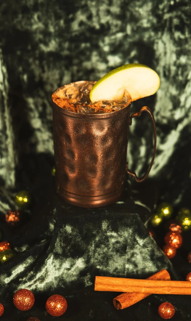Hammered copper mug with dark drink, apple, cinnamon on green velvet.