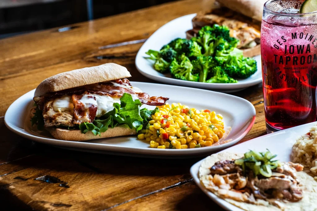 Ciabatta sandwich, chicken, broccoli, taco, and drink on rustic wooden table.