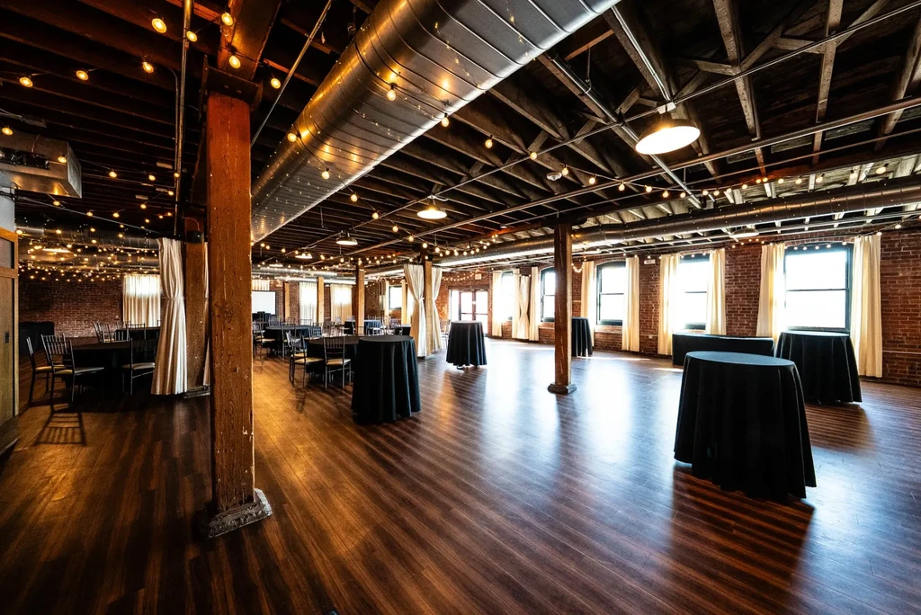 Empty industrial-chic event space with brick walls, tables, and warm lighting.