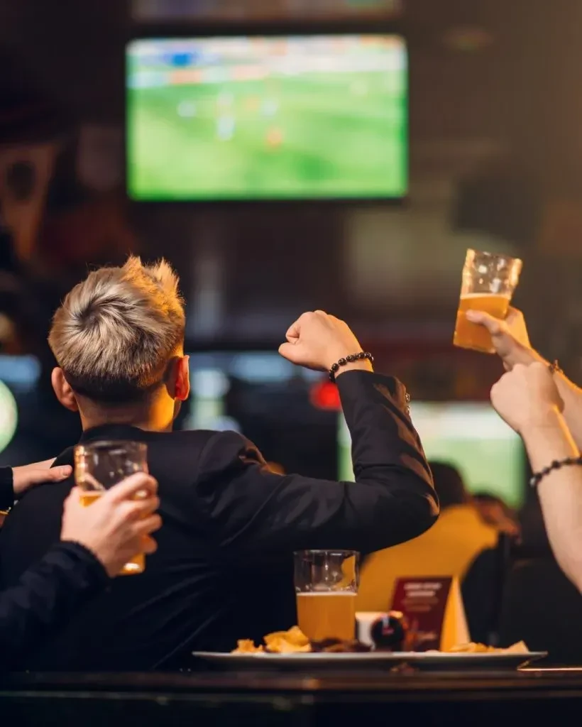 Blonde spiky-haired person cheers with beer watching sports on TV.
