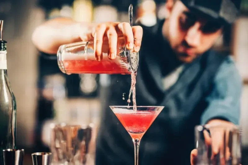 A bartender strains a red cocktail into a martini glass.