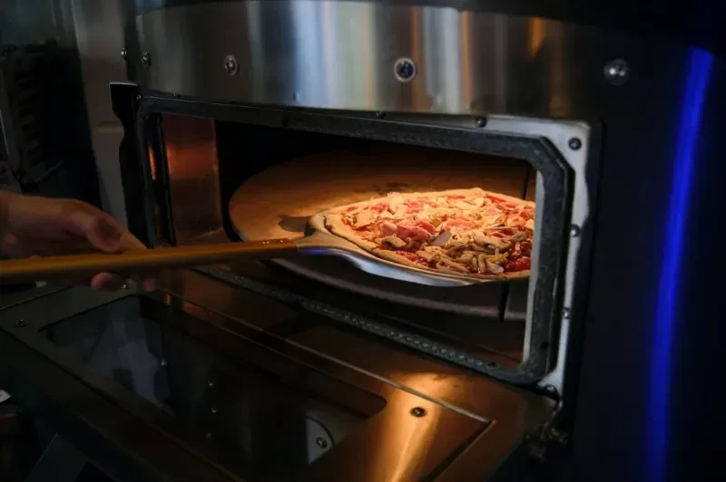 Raw pizza with ham and cheese slides into a glowing oven.
