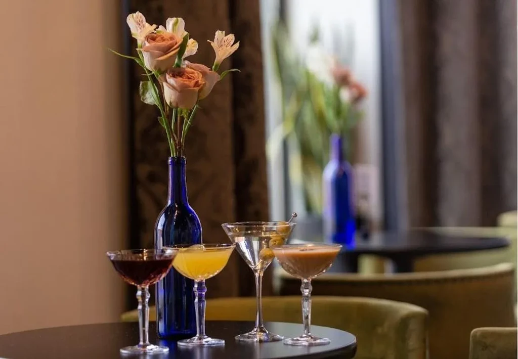 Four colorful cocktails surround a rose bouquet on a dark table.