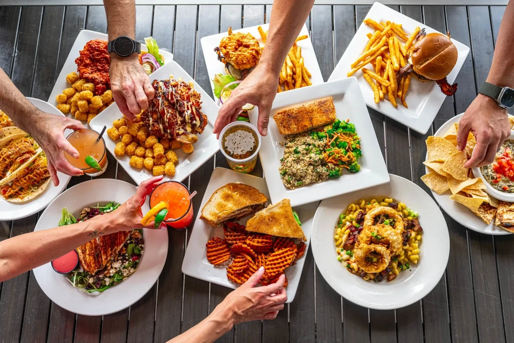 Hands reach for varied dishes and drinks on a dark wooden table.