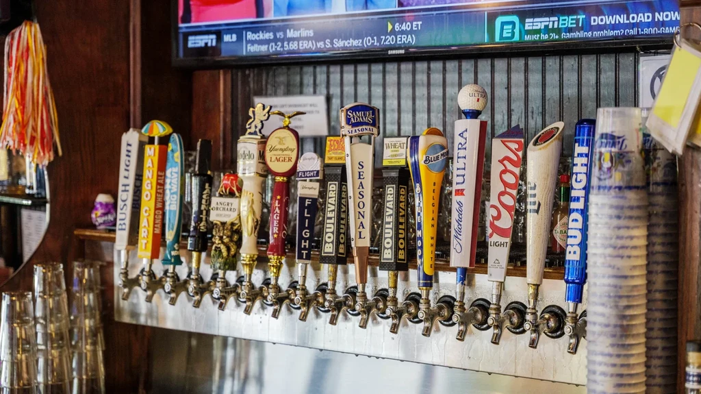 Row of various beer tap handles at a bar, showing sports TV.