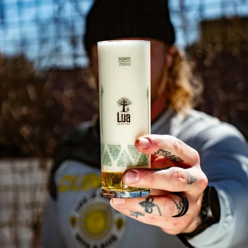 Tattooed hand holds branded Lua Brewing glass with golden foamy beer.