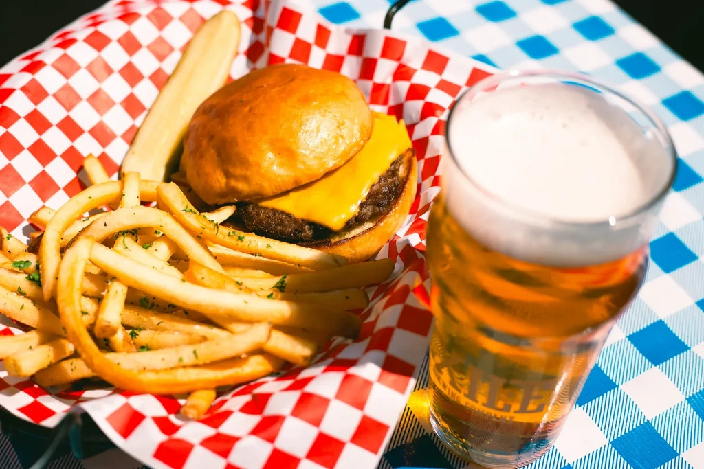 Top-down view of cheeseburger, fries, pickle, beer on checkered tablecloth.