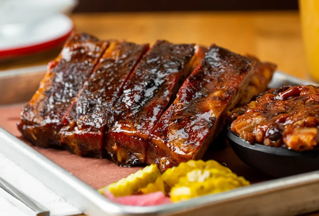 Close-up of sauced barbecue ribs, baked beans, and pickles on a tray.