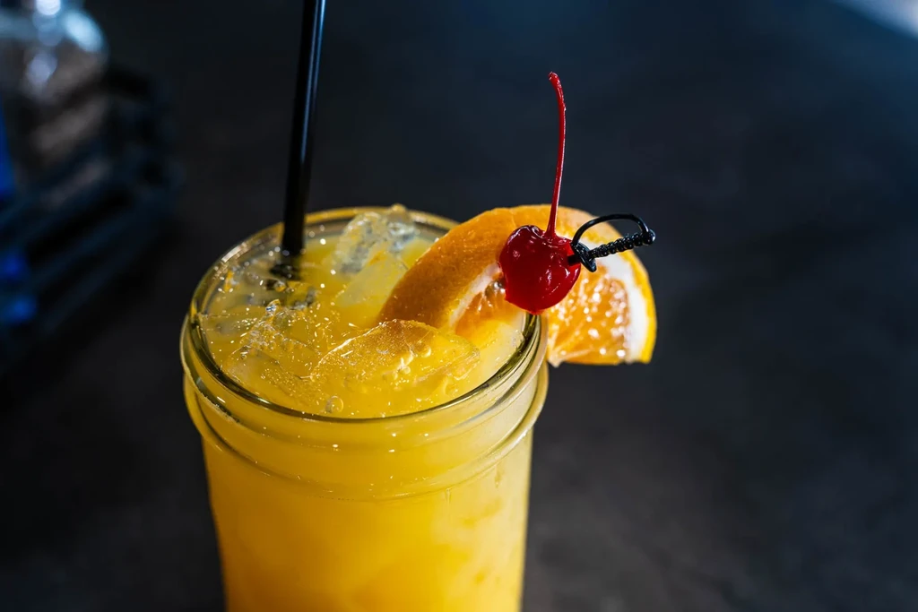 Yellow cocktail with ice, orange slice, cherry on sword pick.