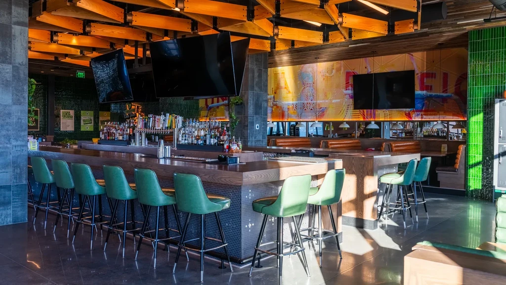 Modern bar with long wooden counter, green stools, and geometric ceiling.