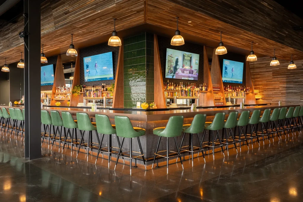 Empty modern L-shaped bar with green stools, liquor display, and multiple TVs.