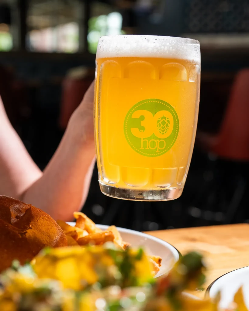Close-up of frothy amber beer in '30 hop' mug, with blurry food.