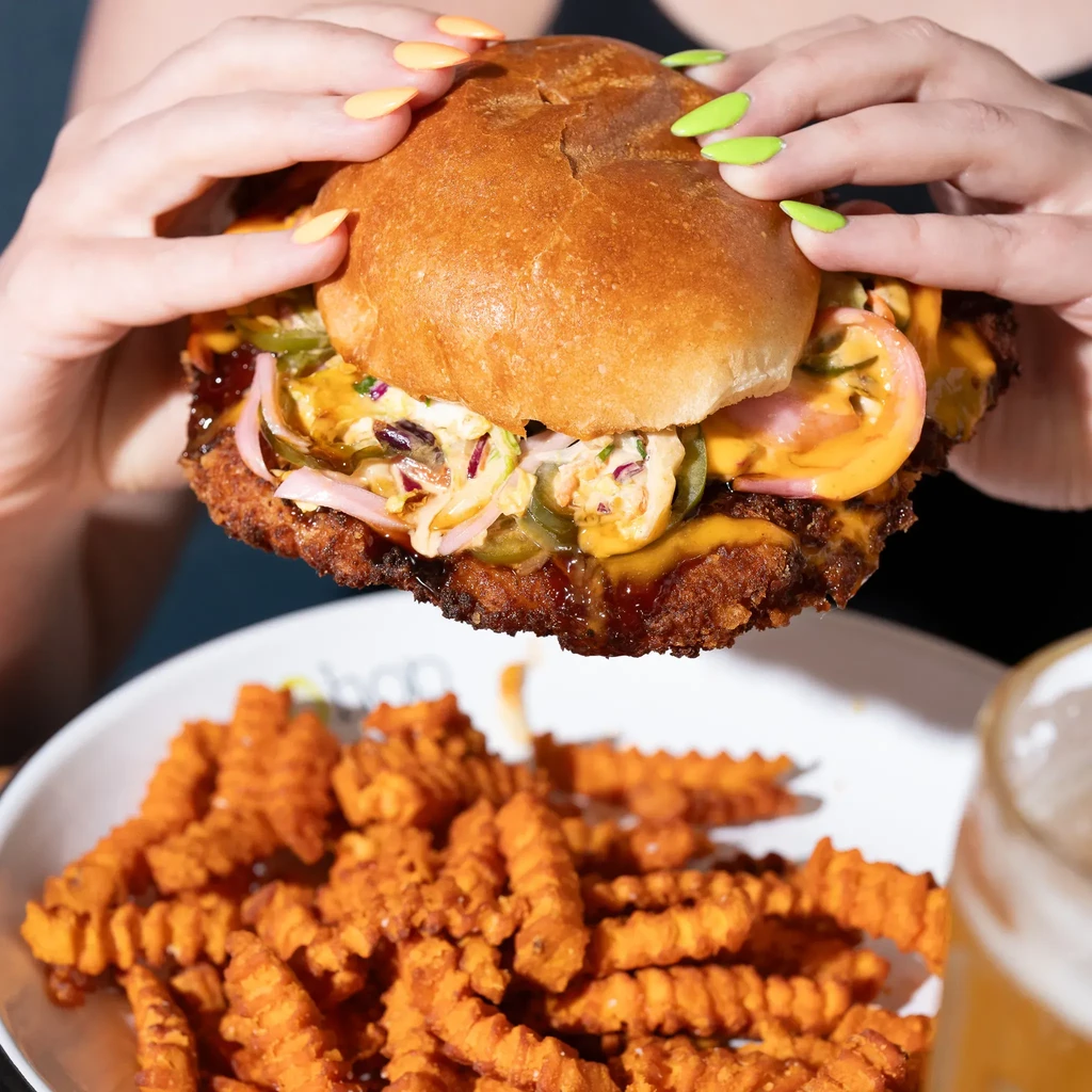 Brightly-nailed hands hold a messy fried chicken sandwich with sweet potato fries.